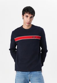 Mornarsko rebrasto dolg rokav T-shirt v temno modri barvi z rdečim in belim vodoravnim pasom preko prsnega dela, z majhno oznako logotipa.