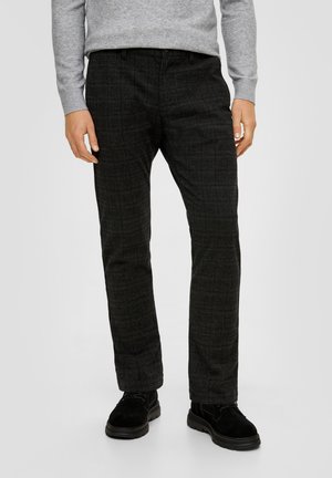 Homme portant un pantalon à carreaux gris foncé, des chaussures noires et un pull gris clair, debout devant un fond uni clair.