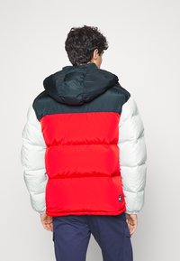 Chaqueta acolchada roja y blanca con capucha verde oscuro. Textura acolchada con un relleno prominente y detalle del logo en la parte inferior izquierda. Capucha ajustable.