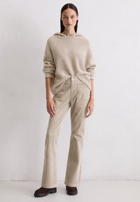 Maglione cappuccio beige e morbido con maniche arrotolate; pantaloni a palazzo in velluto a coste leggeri di colore nocciola con bottoni e tasche frontali; scarpe con zeppa marrone scuro.