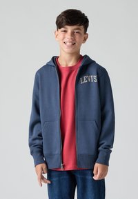 Felpa con cappuccio blu navy con zip e due tasche frontali, logo "LEVI'S" in beige sulla parte superiore sinistra. Indossata sopra una maglietta rossa e jeans blu.