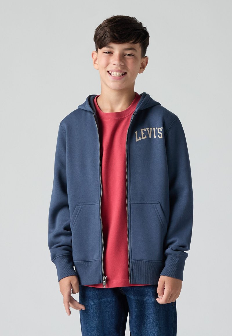 Felpa con cappuccio blu navy con zip e due tasche frontali, logo "LEVI'S" in beige sulla parte superiore sinistra. Indossata sopra una maglietta rossa e jeans blu.
