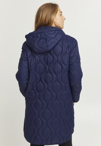 Cappotto trapuntato blu navy con cappuccio, caratterizzato da un motivo testurizzato di forme ovali sovrapposte e un design di lunghezza media.