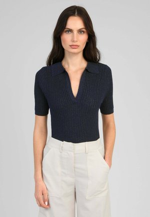 Femme aux longs cheveux foncés portant un haut côtelé bleu marine à manches courtes avec col et décolleté en V profond, associé à un pantalon taille haute beige clair.