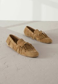 Scarpe mocassino in suede beige con dettagli in frangia, accenti in pizzo e occhielli dorati. Texture morbida e design a punta arrotondata.
