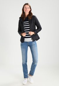 Casaco preto acolchoado com fecho, suéter listrado em branco e azul marinho por baixo, jeans azuis e ténis brancos. Tecido suave, design ajustado.