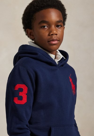 Junge trägt einen marineblauen Hoodie mit einer roten Drei auf dem Ärmel und einem roten Polo-Spieler-Logo auf der Brust.