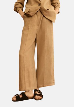 Persona che indossa pantaloni beige a gamba larga e giacca abbinata con le mani nelle tasche, abbinata a sandali neri con fibbia.