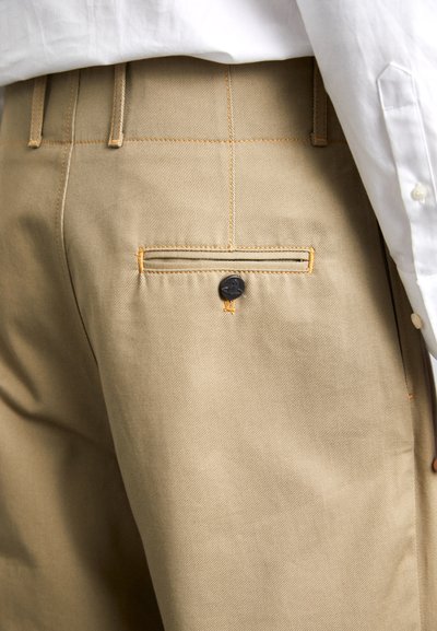 Beige Baumwollhosen mit einem subtilen Fischgrätmuster. Eigenschaften sind orangefarbene Nähte und eine Gesäßtasche mit einem schwarzen Knopfverschluss.