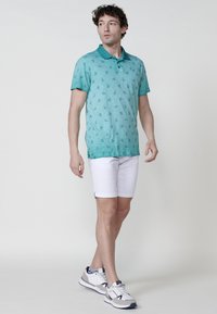 Türkiz színű pólóing finom levélmintával, rövid ujjakkal és gallérral; fehér felgöngyölt rövidnadrággal és szürke sportcipővel párosítva.