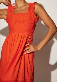 Femme portant une robe orange sans manches avec des bretelles volantées et des motifs de palmiers brodés sur le corsage, main sur la hanche.