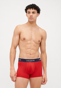 Boxers slip vermelhos feitos de tecido elástico com um cós azul-marinho com o logótipo "LACOSTE". Textura suave, design ajustado e comprimento até à metade da coxa.