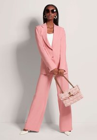 Roze op maat gemaakte blazer en wijde broek, gecombineerd met een gestructureerde lichtroze handtas en witte schoenen met puntige neus. Overmaatse zonnebril.