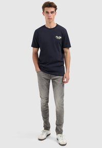 Jonge man staand in een marineblauw graphic t-shirt, grijze slim jeans en witte sneakers tegen een effen lichte achtergrond.