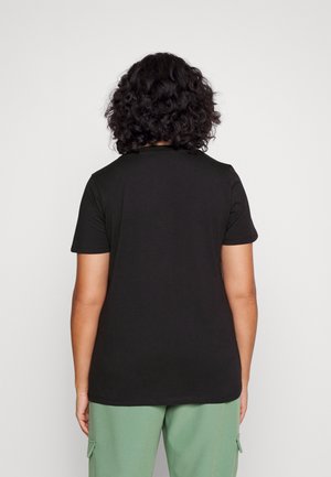 Persoon met krullend haar staat met de rug naar de camera, draagt een effen zwart T-shirt met korte mouwen en lichtgroene broek tegen een witte achtergrond.