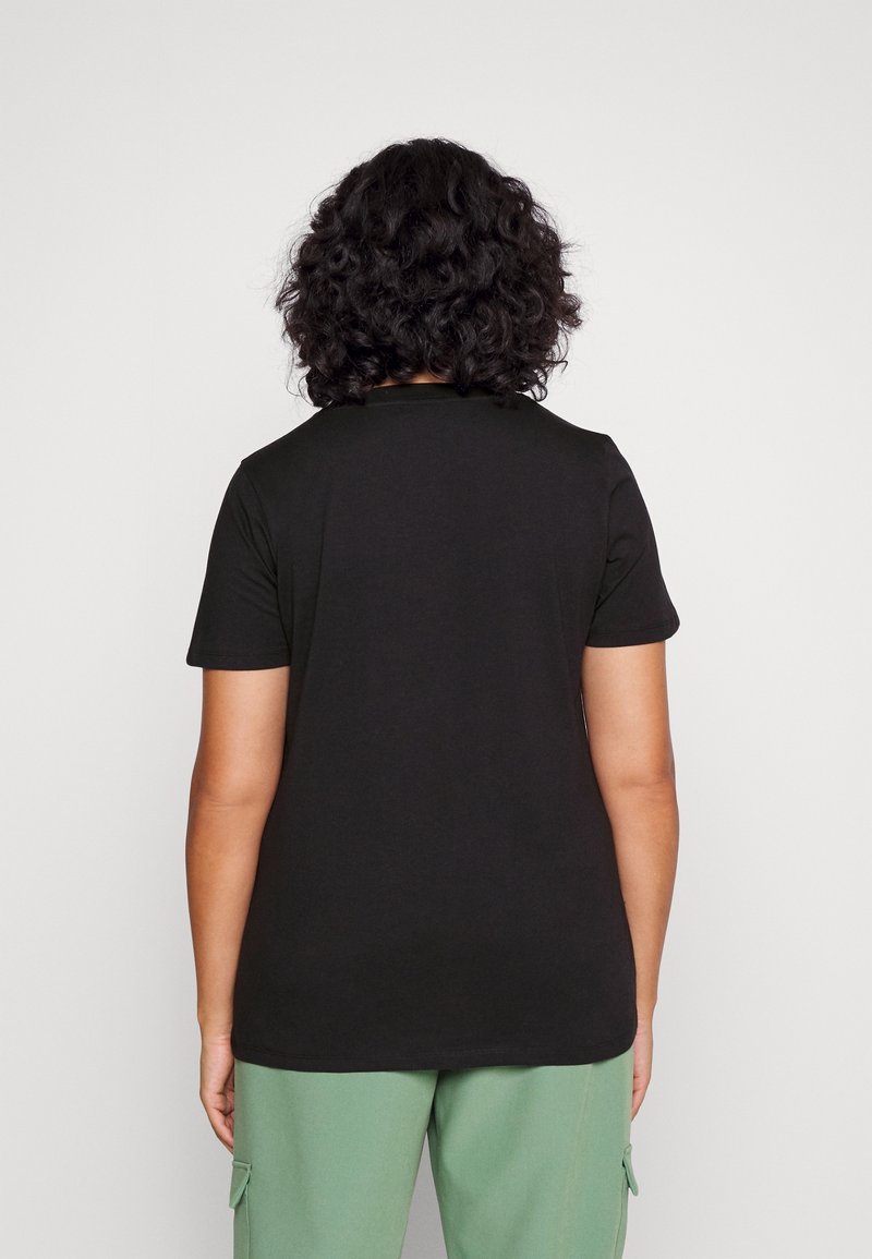 Personne aux cheveux bouclés vue de dos, portant un t-shirt noir uni à manches courtes et un pantalon vert clair sur un fond blanc.