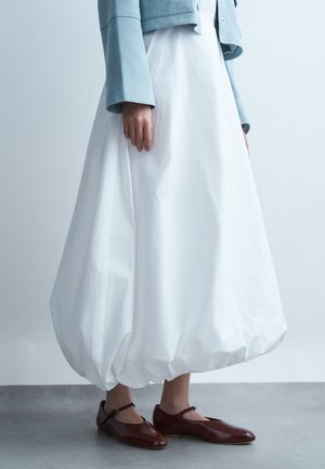 Personne portant une jupe ballon blanche, une veste courte bleu clair et des ballerines Mary Jane marron, debout sur un sol gris contre un mur pâle.