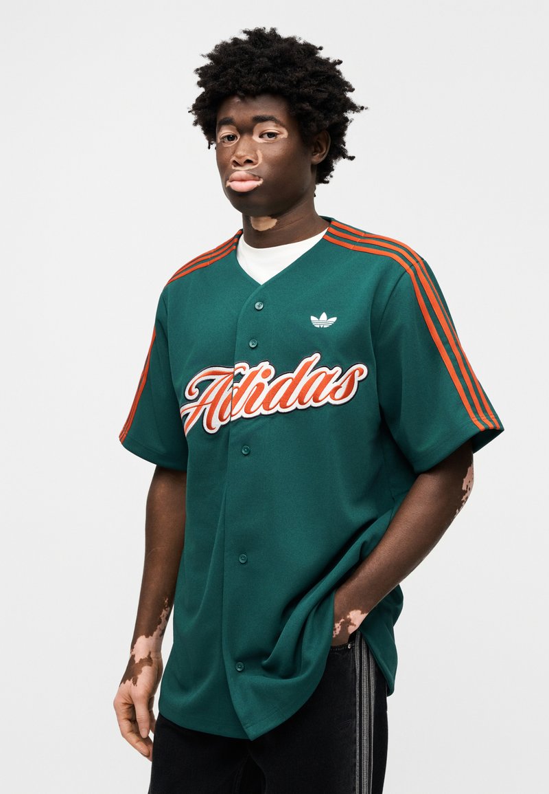 Maillot vert avec le lettrage "Adidas" orange, manches courtes, et trois bandes orange sur les épaules. Associez-le à un pantalon foncé pour un contraste.