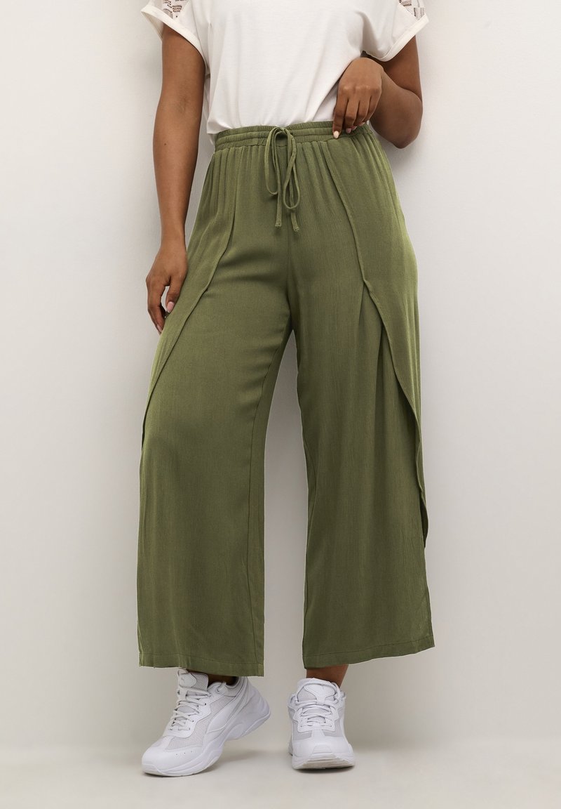 Pantalon vert à jambes larges avec fentes latérales, taille à cordon, tissu texturé, associé à des baskets blanches.