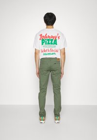 T-shirt blanc avec du texte vert et rouge "Pizza de Johnny" et graphique. Pantalon olive. Personne de dos, portant des baskets beiges.