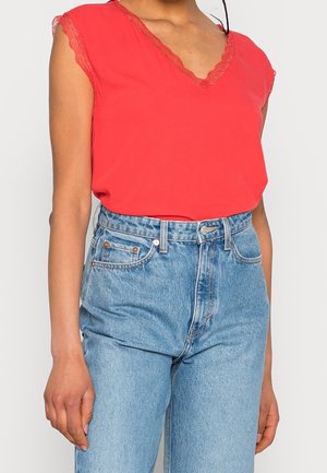 Femme portant un haut rouge sans manches avec une bordure en dentelle et un jean bleu clair taille haute, debout devant un fond clair uni.