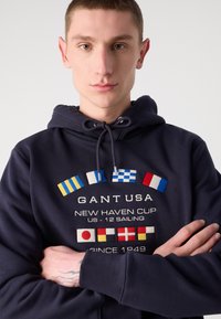 Felpa navy con cappuccio, caratterizzata da bandiere nautiche ricamate, logo frontale e cordino. Texture morbida con polsini e orlo a coste.