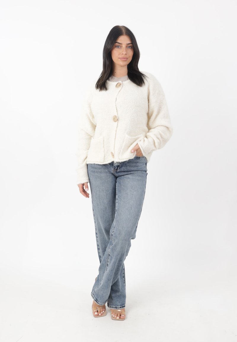 Cardigan fluffy color crema con scollo rotondo, tre grandi bottoni in legno e due tasche frontali. Indossato sopra dei jeans blu.