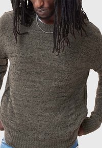 Dark olive green knitted sweater with a textured finish, round neckline, and ribbed hem. Worn over a white shirt, featuring long sleeves.