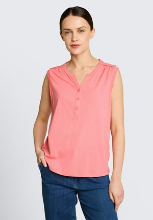 Kobieta o ciemnych włosach, ubrana w różową bezrękawnikową bluzkę na guziki i niebieskie dżinsy, stojąca z jedną ręką w kieszeni na jednolitym tle.