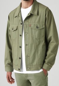 Chaqueta de mezclilla verde de algodón con bolsillos delanteros, botones negros y un corte ligeramente cropped. Combinada con una camiseta blanca debajo.