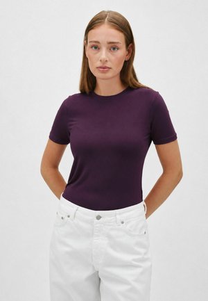 Donna con capelli castani lisci che indossa una maglietta viola a maniche corte e pantaloni bianchi, in piedi davanti a uno sfondo chiaro e semplice.