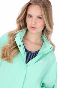 Chaqueta ligera verde menta con cuello alto, botones de presión y un logo sutil. Llevada sobre una camisa oscura con rayas. Textura suave.