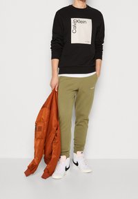 Felpa nera con logo Calvin Klein, pantaloni jogger verde oliva, giacca bomber arancione e sneakers bianche alte. Tessuto liscia e vestibilità rilassata.