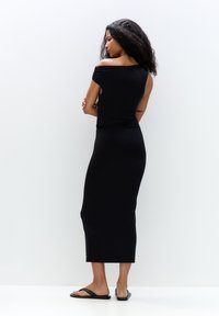 Schwarzes, eng anliegendes Maxikleid mit schulterfreiem Design, glatter Textur und einem Oberschenkelspalt hinten. Kombiniert mit flachen schwarzen Sandalen.