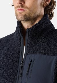 Giacca in pile di colore blu navy, con parte superiore in pelliccia testurizzata e parte inferiore in tessuto liscio. Presenta una chiusura con zip e dettagli con logo sulla tasca del petto.