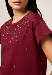 T-shirt court en maroon à manches courtes avec un col rond, présentant des embellissements en strass dispersés de différentes tailles sur le devant.