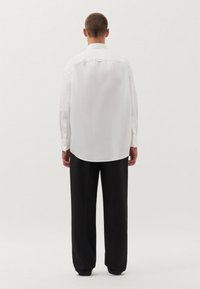 Witte blouse met lange mouwen en een ronde onderrand, zichtbare knoopdetails bij de mouwen, gecombineerd met losse zwarte broek, gezien vanaf de achterkant.