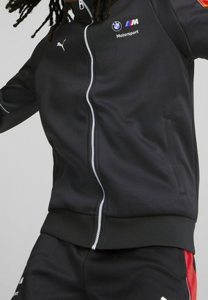 Veste noire zippée en tissu lisse, arborant les logos BMW et Puma, des accents blancs et des poches. Associée à un pantalon de jogging noir.