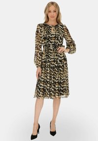 Czarna i beżowa sukienka z długimi przezroczystymi rękawami, teksturowany wzór w zygzaki, ściągnięta w talii paskiem, długość do kolan, w połączeniu z czarnymi szpilkami.