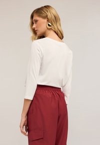 Vaalea nainen, jolla on aaltoileva hius, pukeutuneena suuriin kultaketjuihin, valkoiseen pitkähihaiseen paitaan ja korkeavyötäröisiin burgundinvärisiin housuihin, joissa on sivutaskut, katsoo vasemmalle.