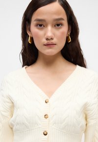 Cardigan crem cu tricotaj de tip cable-knit, cu decolteu în V, textură ribbită și trei nasturi mari în față. Accentuat cu cercei aurii în formă de cerc.