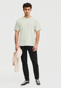 Jasnozielona bawełniana koszulka, krótki rękaw, w zestawie z czarnymi spodniami slim-fit. Kremowy sweatshirt zarzucony na lewy ramie, na stopach czarne trampki.