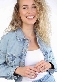 Denim jas met zilveren knopen, lichtblauwe kleur, gedragen over een witte top. Model draagt twee ringen, die textuurdetails op de vingers tonen.