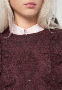 Burgundowa dzianinowa sweter z teksturowymi detalami, z okrągłym dekoltem oraz jasnoróżową koszulą z kołnierzykiem pod spodem, z zapięciem na guziki.