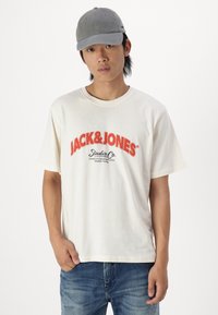 Camiseta de algodón crema con texto y logo rojos de "JACK&JONES". Manga corta, escote redondo, combinada con jeans de mezclilla azules y una gorra gris.