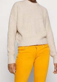 Pullover beige a maglia con scollatura rotonda e maniche a costine, abbinato a pantaloni in velluto a coste giallo brillante. La texture appare morbida e accogliente.