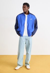 Nike vējjaka zilā un tumši zilā krāsā, ar rāvējslēdzēju, stāvošu apkakli un logotipu. Saderēta ar gaiši zilām džinsiem un baltām kedām ar gumijas zole.