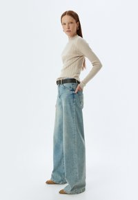 Beżowa, prążkowana bluzka z długim rękawem, luźne jasnoniebieskie jeansy z szerokimi nogawkami, czarny pasek i beżowe obuwie; modelka stoi z jedną ręką na biodrze.