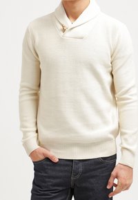 Biały sweter dzianinowy z kołnierzem w stylu szala, z drewnianym zapięciem na guziki. Ściągacze na mankietach i u dołu, noszony z ciemnymi dżinsami. Gładka faktura.