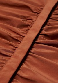 Stof in roestkleur met een gladde textuur, voorzien van verticale plooien en een platte strook in het midden, die de structuur van het kledingstuk benadrukt.